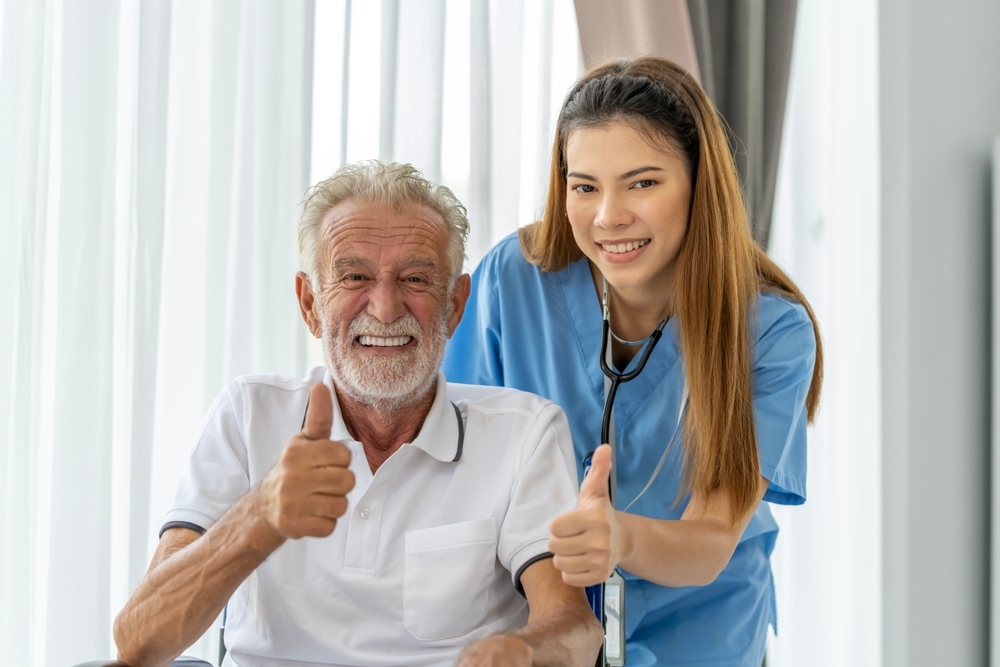 Smiling female nurse in blue scrubs giving a thumbs up alongside a happy elderly male patient, representing the compassionate care provided to patients who qualifies for private duty nursing