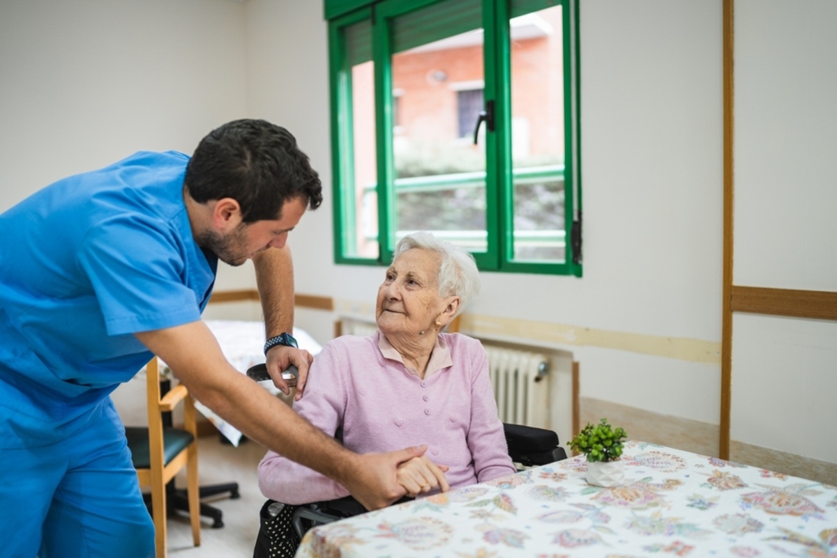 Male nurse in blue scrubs assisting an elderly female patient, representing LPN to RN bridge program through hands-on home care experience