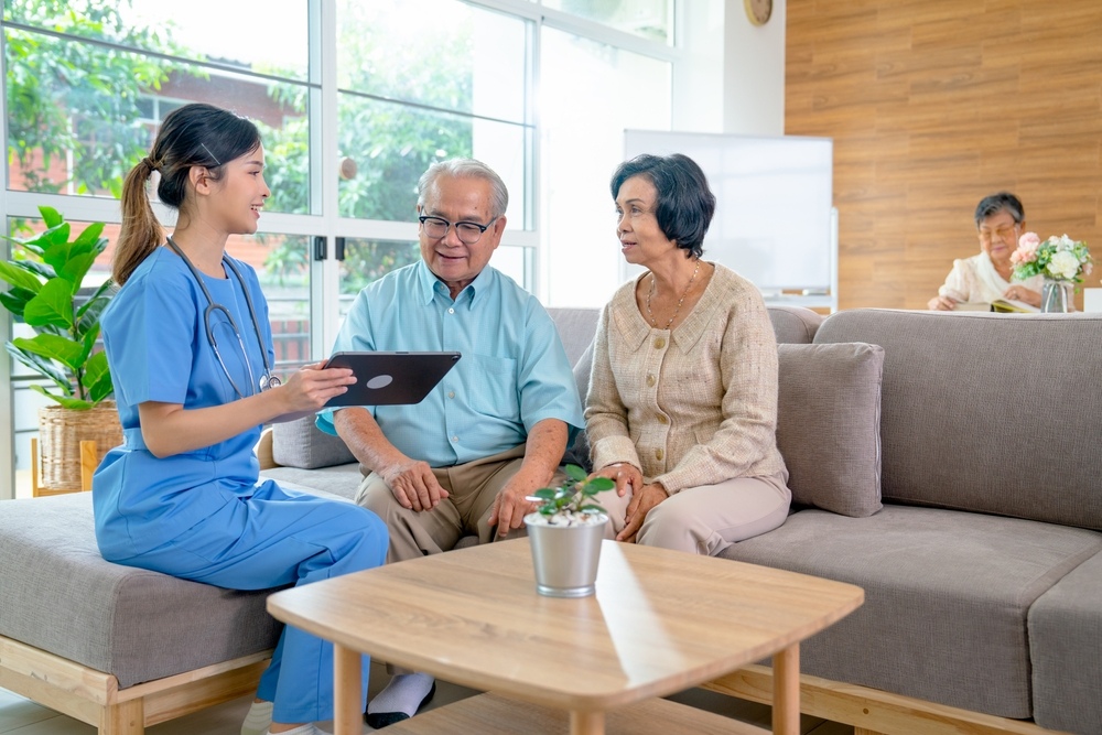 Home care nurse in blue scrubs showing a tablet to an elderly couple on a sofa, representing the personalized care coordination provided by a private duty nursing agency