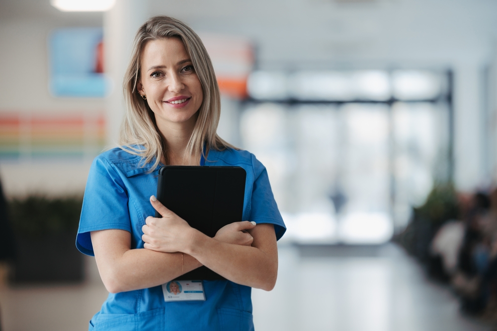 Smiling female nurse in teal scrubs holding a tablet with a name badge, representing the professional competence behind a competitive private duty nurse salary in New York