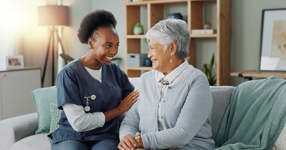 Private duty nurse jobs in Queens NY - smiling RN in navy scrubs sitting with elderly female patient on couch during in-home care visit