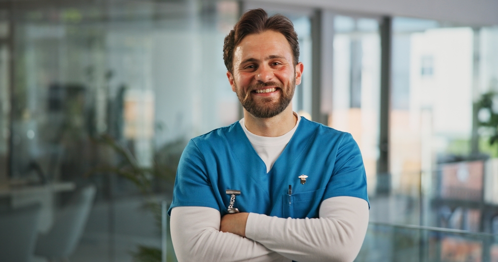 Smiling male nurse in blue scrubs standing with arms crossed in a clinical hallway, representing the confidence and fulfillment that come from the most rewarding parts of being an LPN